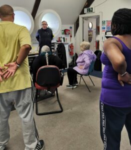 an exercise instructor demonstrates a chest stretch to 2 seated and 2 standing participants in the gym